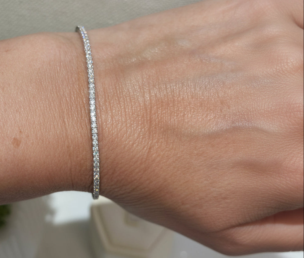 A person wearing a white watch is showcasing a silver bangle bracelet adorned with cubic zirconia stones on their wrist.